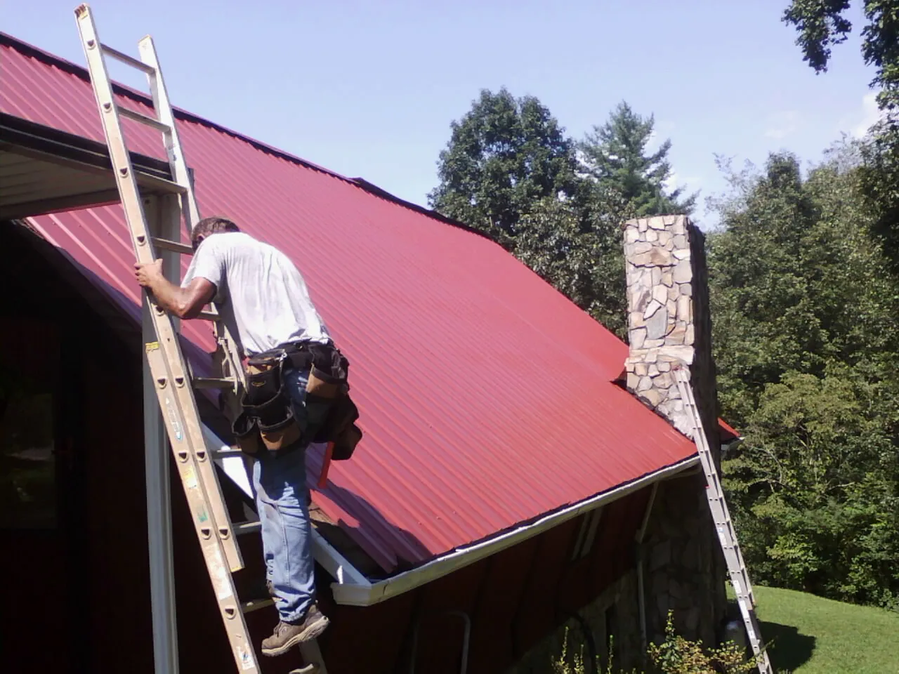 Roofer on a ladder beside a red metal roof and stone chimney — Soffit & Fascia Repair by Roofing Brothers in Sproul